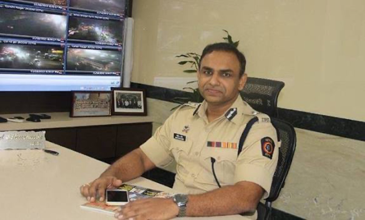 Milind Bharambe in uniform seated at a desk in a police control room, with a bank of traffic camera monitors on the wall behind him.