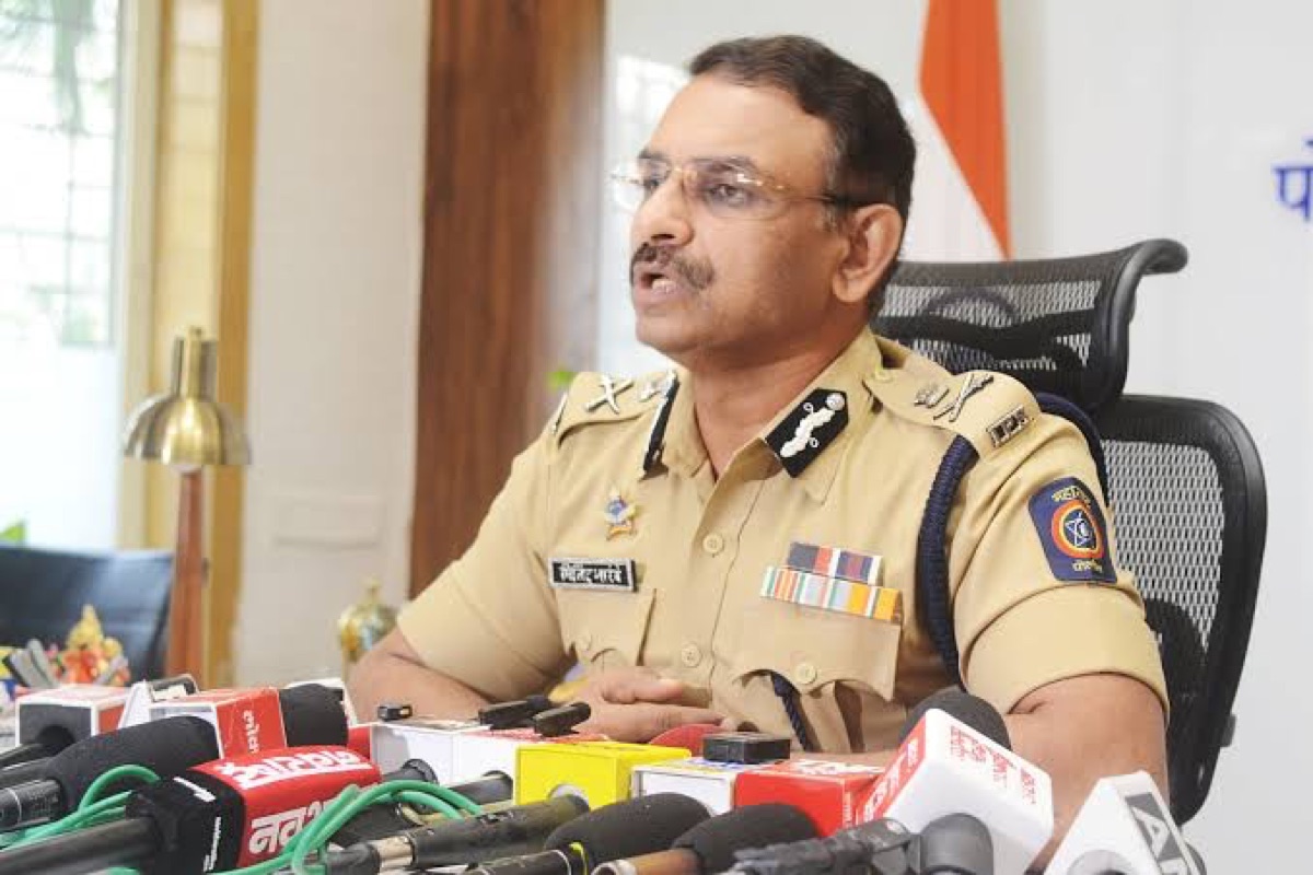 Milind Bharambe addressing a press briefing at a table of assembled microphones, speaking to reporters.