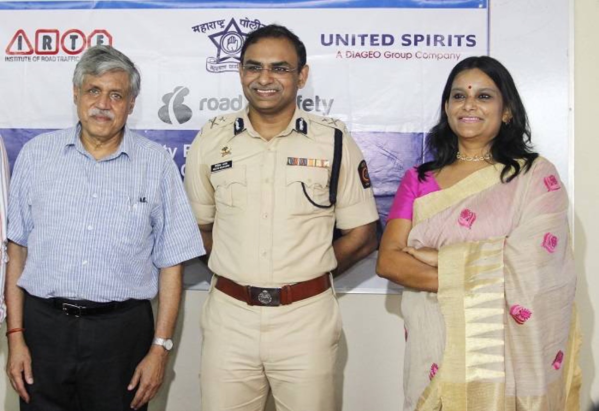Milind Bharambe in uniform standing with two other guests at a road safety event, before a backdrop naming the Institute of Road Traffic Education and a road safety programme.