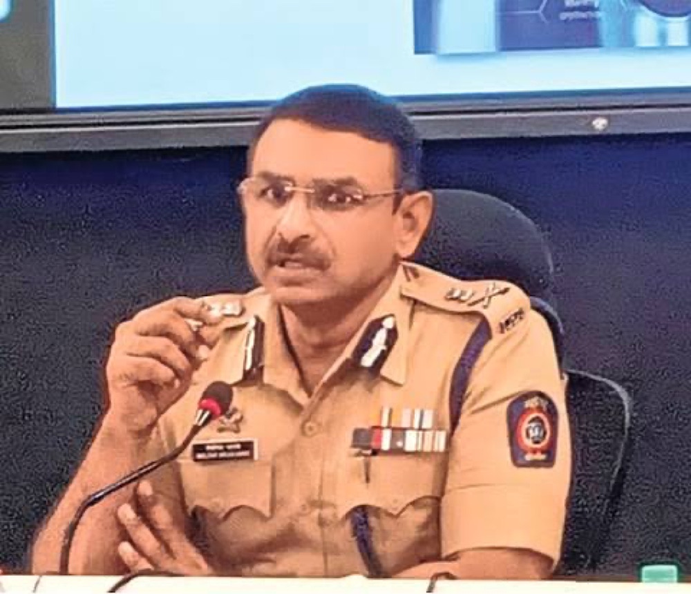 Milind Bharambe in uniform speaking at a briefing table with a microphone and a presentation screen behind him.