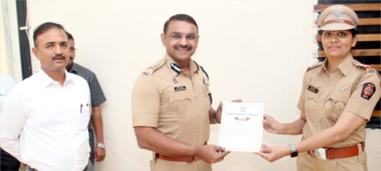 Milind Bharambe in uniform presenting a framed commendation certificate to a woman police officer, with two other people standing alongside.